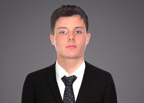 Portrait of a person with short dark hair, wearing a black suit, white shirt, and a dotted tie, with a neutral expression, against a gray gradient background.