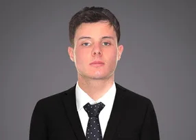 Portrait of a person in a black suit, white shirt and dark polka-dot tie, with neutral expression, short dark hair, against a grey gradient backdrop.