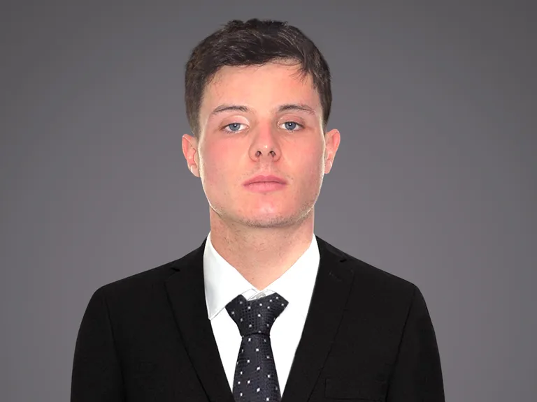 Portrait of a person in a black suit, white shirt and dark polka-dot tie, with neutral expression, short dark hair, against a grey gradient backdrop.