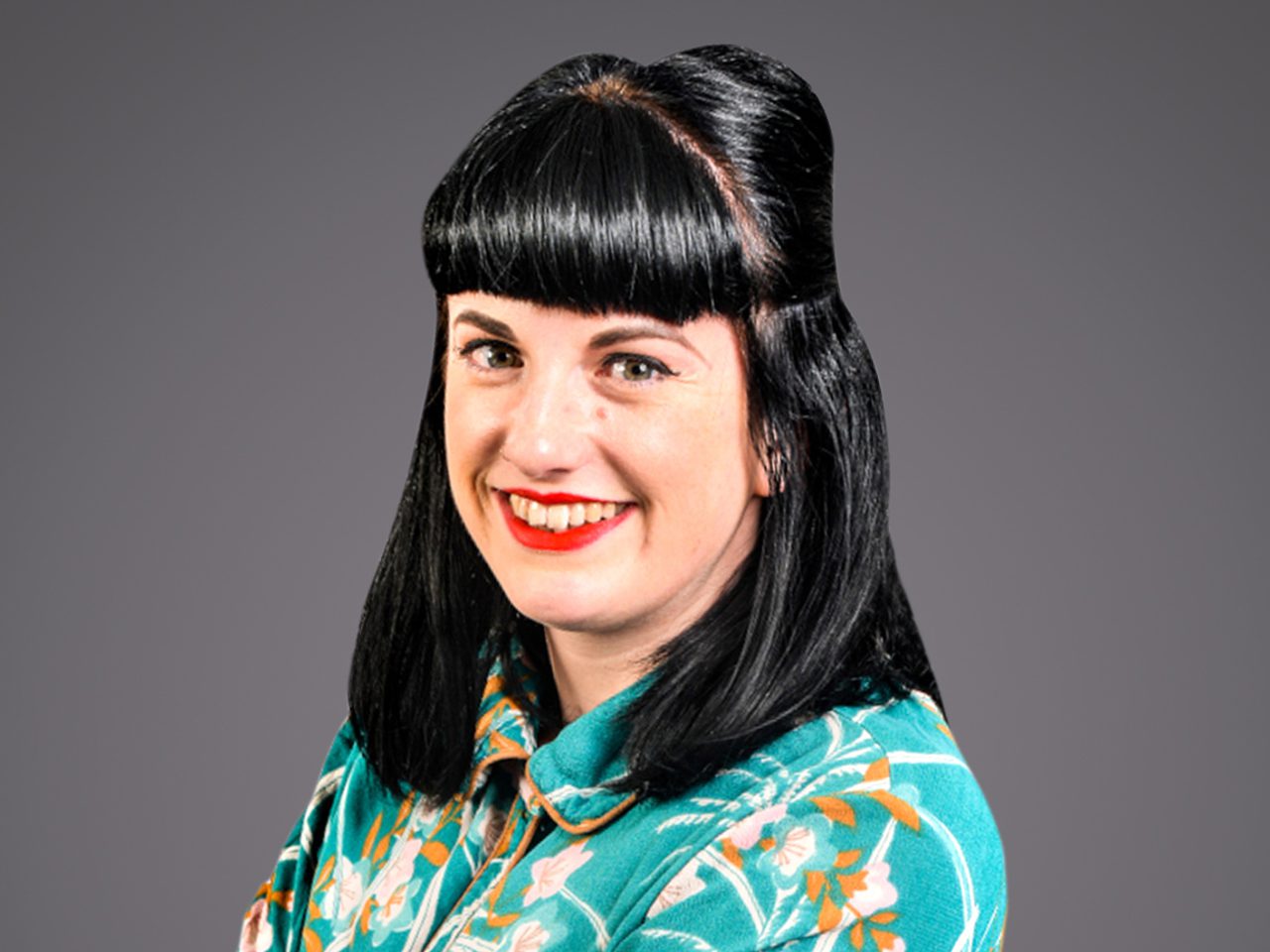 Portrait of a person with black hair in a retro updo and bangs, wearing a teal floral shirt, smiling with red lipstick against a gray backdrop.