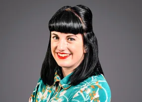 Portrait of a person with straight black hair and blunt bangs, wearing a teal floral shirt, smiling at the camera against a grey studio background.