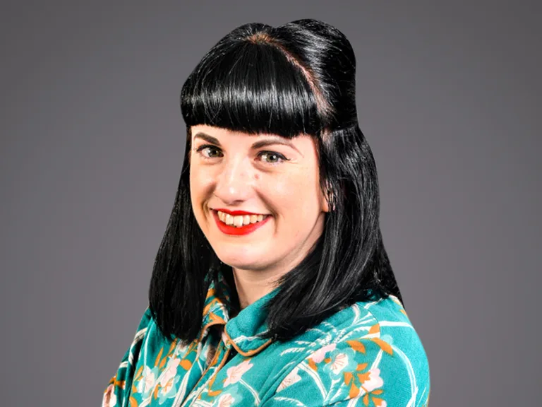 Portrait of a person with straight black hair and blunt bangs, wearing a teal floral shirt, smiling at the camera against a grey studio background.