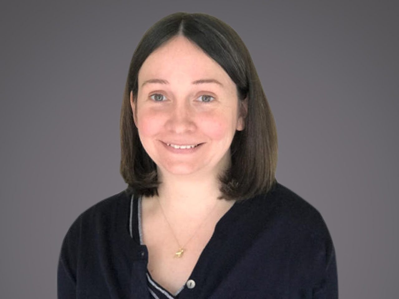 Portrait of a person with shoulder-length brown hair, smiling, wearing a dark cardigan over a striped top, against a neutral grey backdrop.