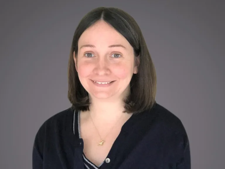 Person with shoulder-length brown hair, smiling, wearing a dark cardigan over a striped top and a gold necklace, against a grey background.