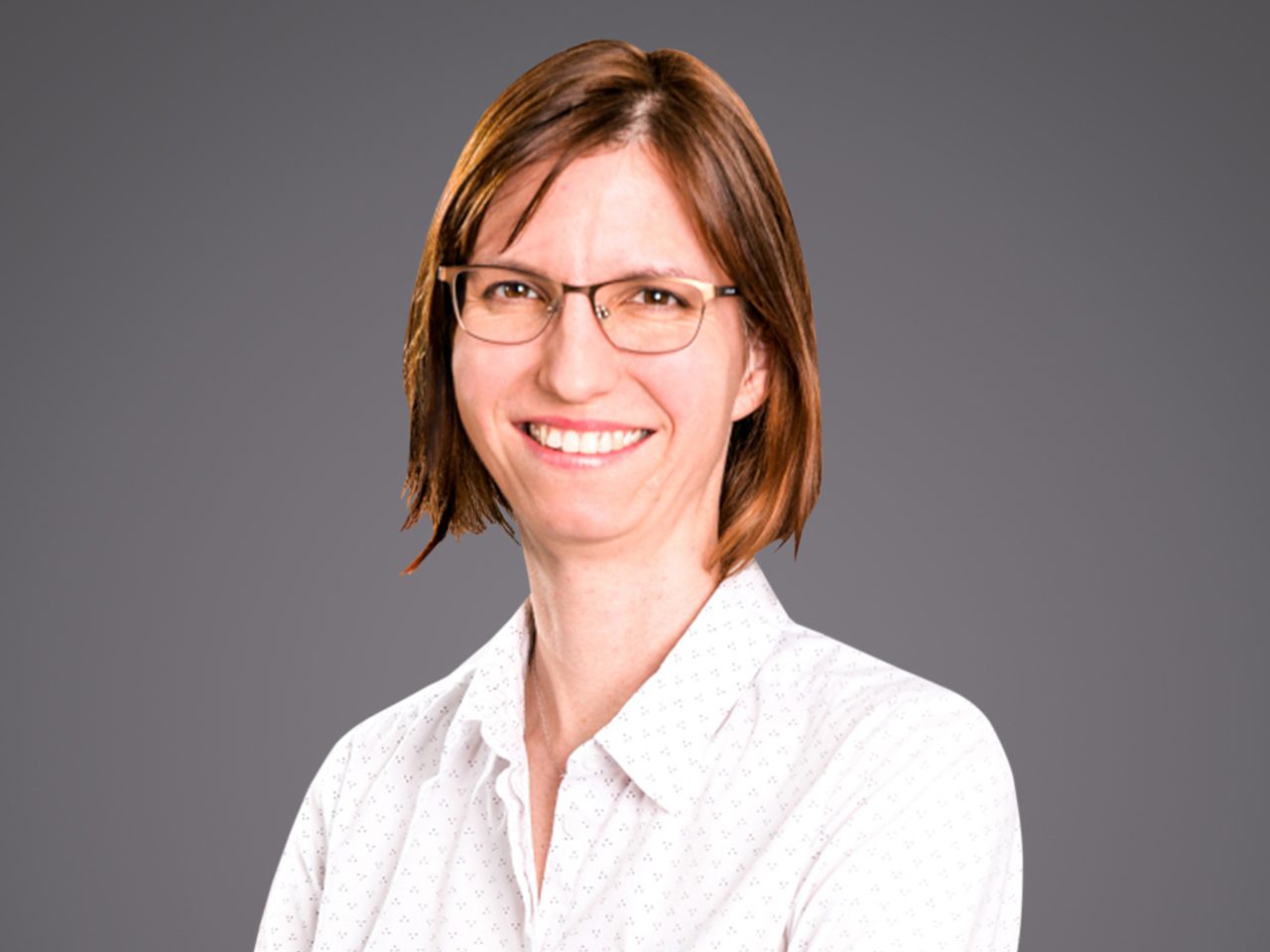 Portrait of a person with short brown hair and glasses, wearing a white dotted shirt, smiling against a grey studio backdrop.