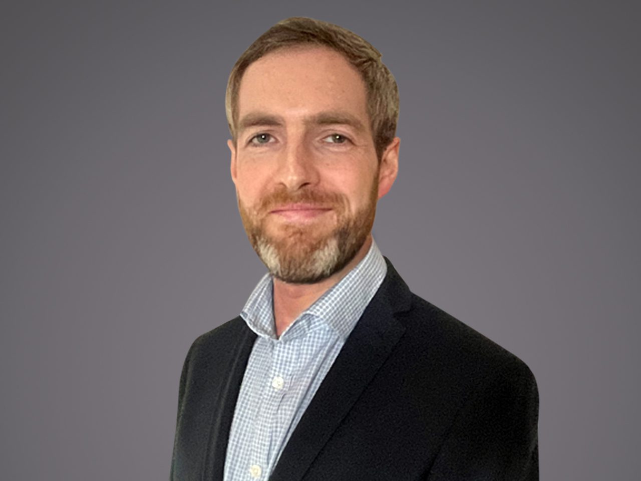 Head-and-shoulders portrait of a person with short light brown hair and beard, wearing a dark blazer over a light check shirt, against a grey background.