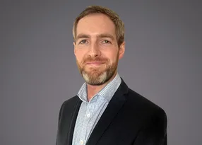 Head-and-shoulders portrait of a person with short light brown hair and beard, wearing a dark blazer over a light check shirt, against a grey background.