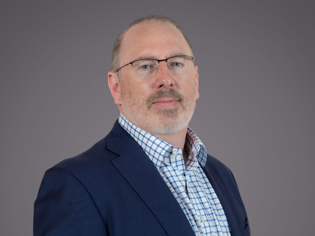 Portrait of a person with short grey hair and glasses, wearing a navy blazer over a blue check shirt, against a neutral grey studio background.