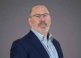 Portrait of a person with short grey hair and glasses, wearing a navy blazer over a blue check shirt, against a neutral grey studio background.