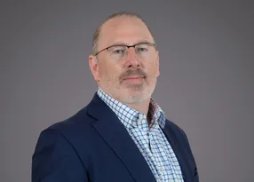 Portrait of a person with short grey hair and glasses, wearing a navy blazer over a blue check shirt, against a neutral grey studio background.