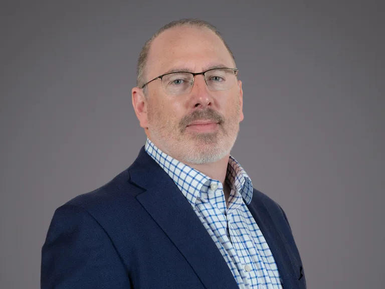 Portrait of a person with short grey hair and glasses, wearing a navy blazer over a blue check shirt, against a neutral grey studio background.