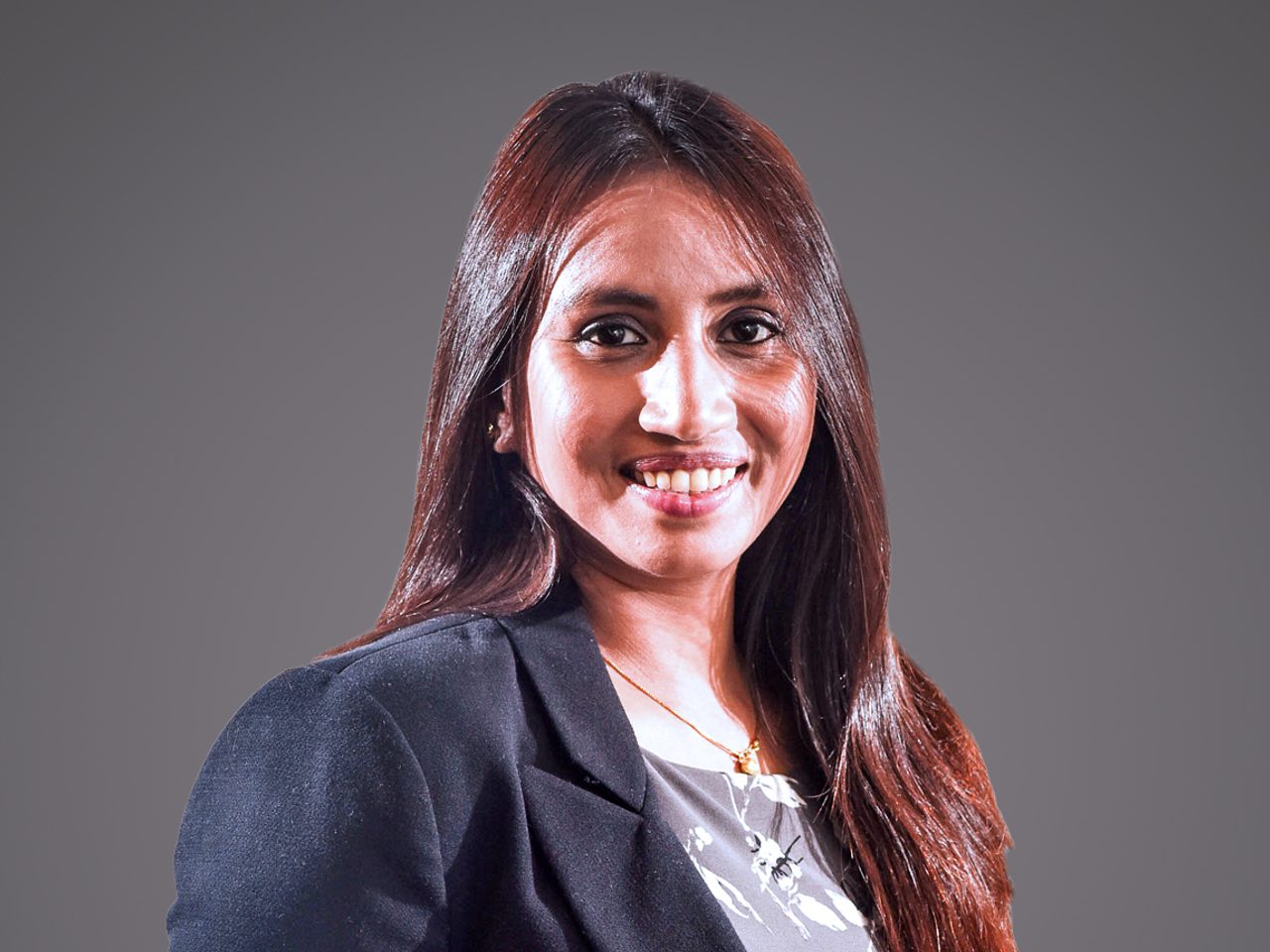Person with long dark hair, wearing a dark blazer and a patterned top, smiling against a neutral gray background.