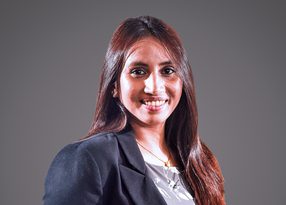 Portrait of a person with long dark hair, wearing a dark blazer over a patterned top, smiling against a grey studio backdrop.