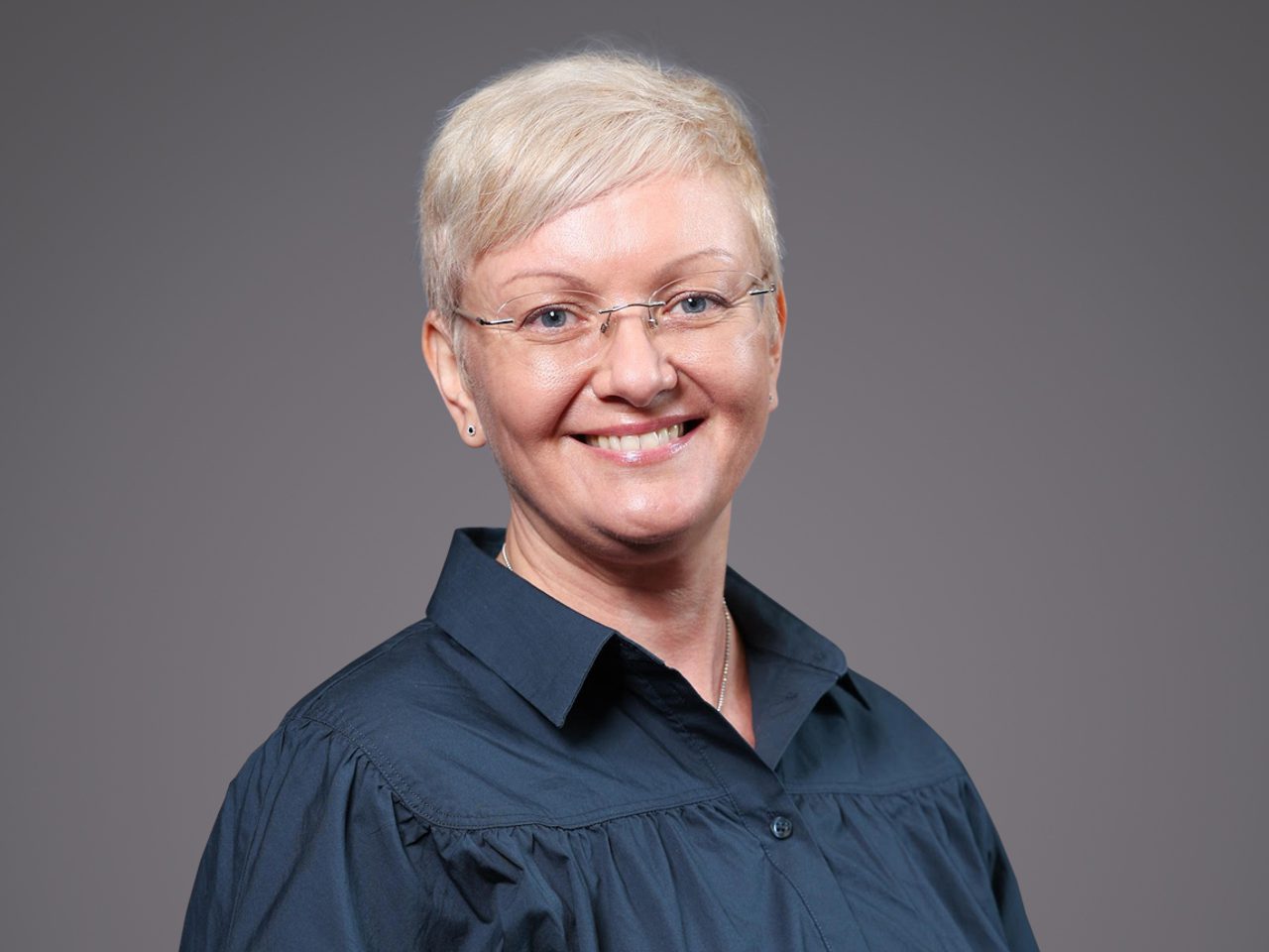 Smiling person with short, light blond hair and glasses, wearing a dark blue collared shirt, posed against a neutral gray backdrop.