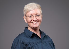 Smiling person with short, light blond hair and glasses, wearing a dark blue collared shirt, posed against a neutral gray backdrop.