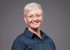 A person with short light blonde hair, wearing wire-rim glasses and a dark blue collared blouse, smiling at the camera against a grey backdrop.