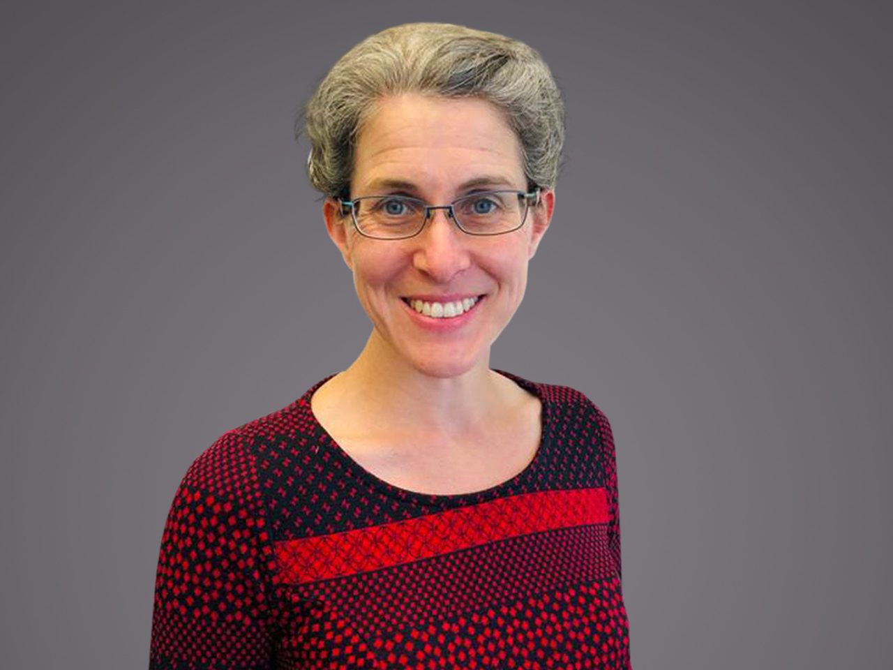 A person with short grey hair and glasses, smiling, wearing a red and black patterned top, against a grey gradient background.
