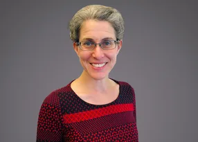 A person with short grey hair and glasses, smiling, wearing a red and black patterned top, against a grey gradient background.