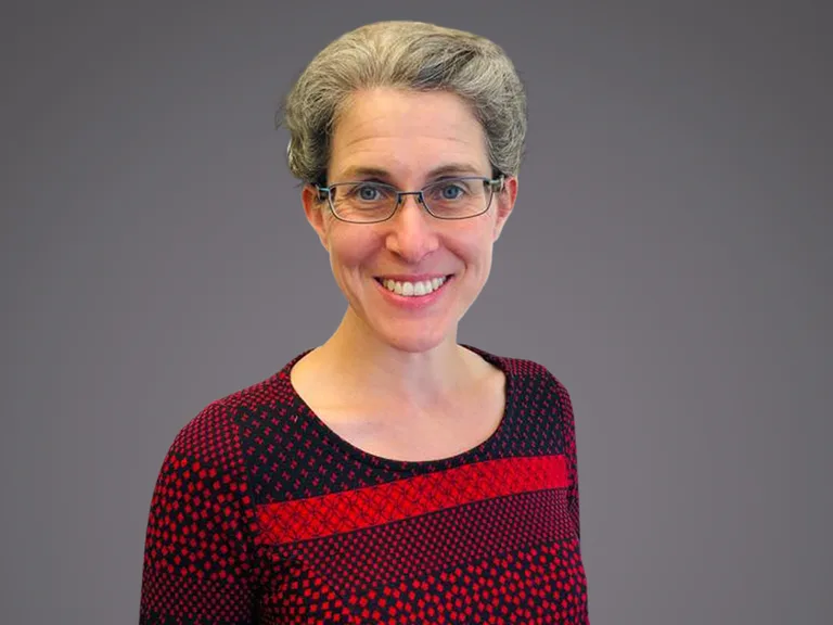 A person with short grey hair and glasses, smiling, wearing a red and black patterned top, against a grey gradient background.