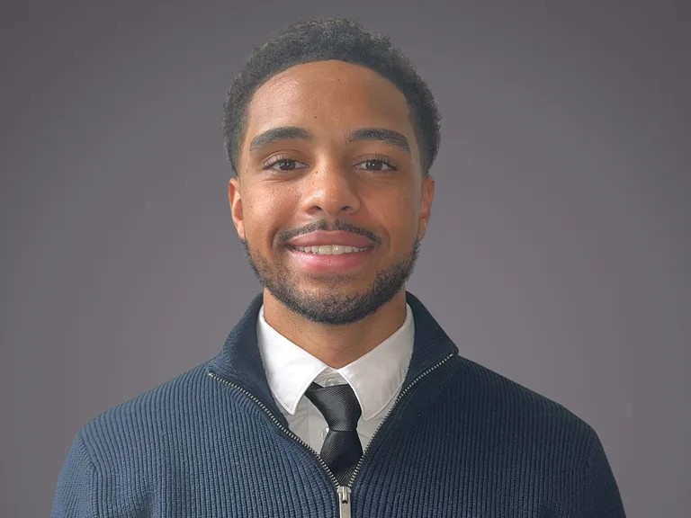 Jeune homme souriant portant une chemise blanche, une cravate noire et un pull zippé, fond gris uni.