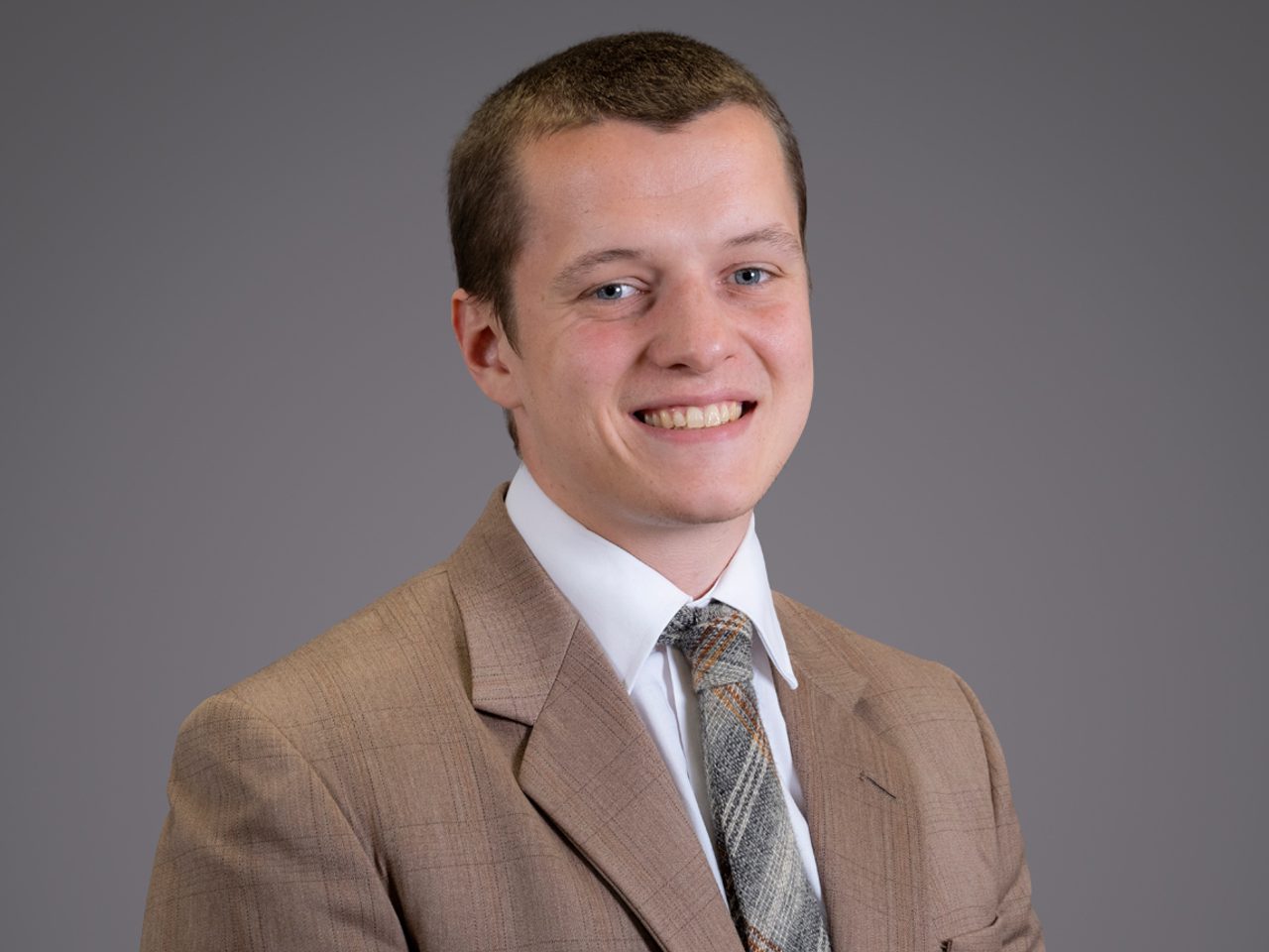 Smiling person in a light brown suit, white shirt, and plaid tie, posed against a plain gray backdrop.
