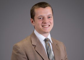 Smiling person in a light brown suit, white shirt, and plaid tie, posed against a plain gray backdrop.