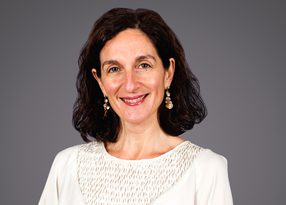 Smiling person with shoulder-length dark hair, wearing a white textured top and dangling earrings, against a neutral grey background.