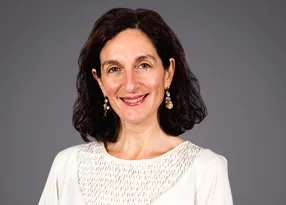 Smiling person with shoulder-length dark hair, wearing a white textured top and dangling earrings, against a neutral grey background.