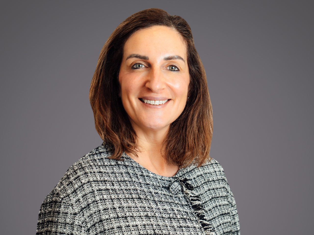 Ritratto di una persona con capelli castani a spalle, sorriso aperto, indossa un top grigio a trama bouclé con un piccolo nodo sul colletto, sfondo grigio.