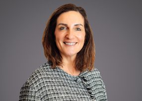 Portrait of a person with shoulder-length brown hair, smiling, wearing a grey-black tweed top with fringe, against a neutral grey background.