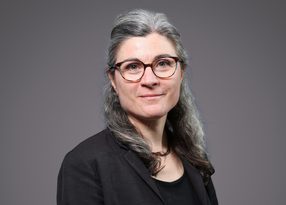 Portrait of a person with long grey hair, wearing round tortoiseshell glasses and a black blazer over a black top, neutral grey backdrop.