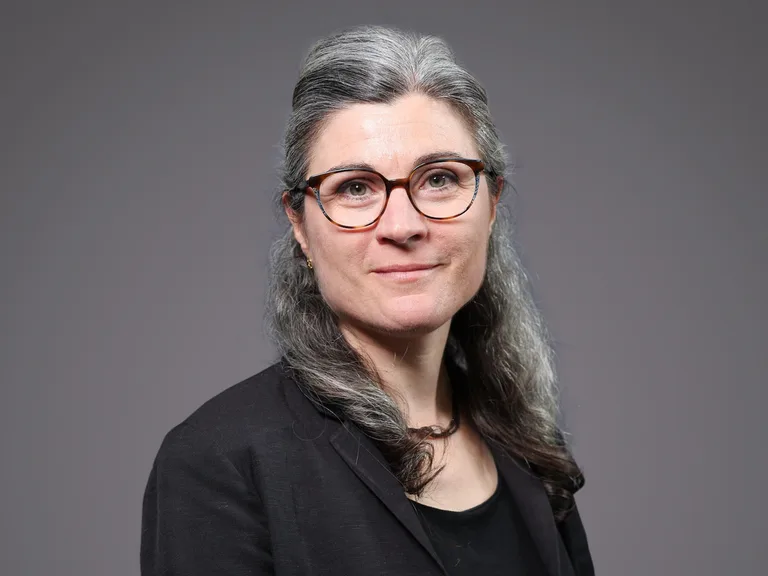 Portrait of a person with long grey hair, wearing round tortoiseshell glasses and a black blazer over a black top, neutral grey backdrop.