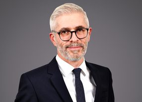 Portrait of a person with short grey hair and a grey beard, wearing black-rimmed glasses, a white shirt, dark suit and tie, against a neutral grey background.