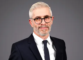 Portrait of a person with short grey hair and a grey beard, wearing black-rimmed glasses, a white shirt, dark suit and tie, against a neutral grey background.