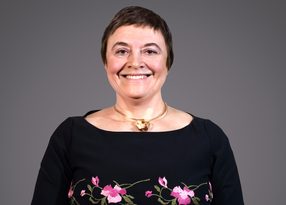 Smiling person with short dark hair, wearing a black top with pink floral embroidery and a gold necklace, against a grey backdrop.