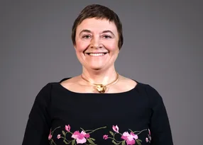 Smiling person with short dark hair, wearing a black top with pink floral embroidery and a gold necklace, against a grey backdrop.