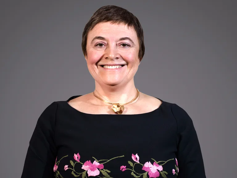 Smiling person with short dark hair, wearing a black top with pink floral embroidery and a gold necklace, against a grey backdrop.