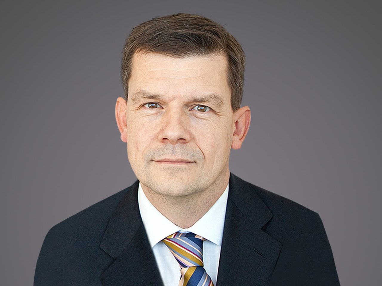 Head-and-shoulders portrait of a person with short brown hair, wearing a dark suit, white shirt and striped tie, against a neutral grey background.