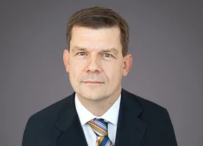 Head-and-shoulders portrait of a person with short brown hair, wearing a dark suit, white shirt and striped tie, against a neutral grey background.