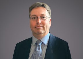 Portrait of a person with short grey hair and glasses, in a dark suit, light shirt and patterned tie, against a neutral grey background.