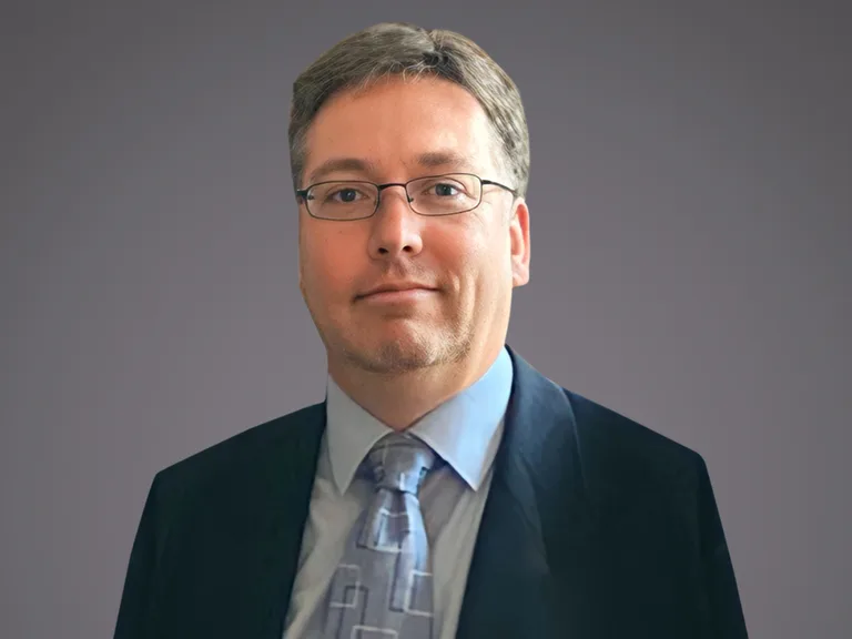 Portrait of a person with short grey hair and glasses, in a dark suit, light shirt and patterned tie, against a neutral grey background.