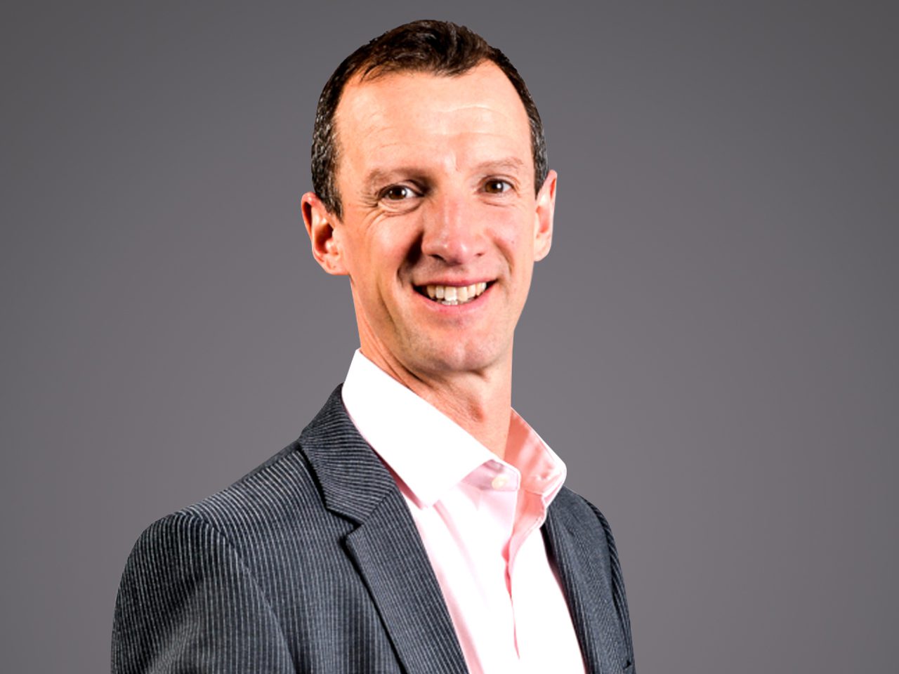 Smiling person in a gray pinstripe blazer and pale pink shirt, facing slightly left, against a neutral gray studio backdrop.