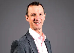 Portrait of a smiling person in a light pink shirt and dark pinstripe blazer, facing slightly to the right, against a neutral grey background.