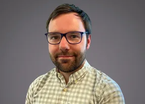 Portrait of a person with short dark hair and a beard, wearing blue-framed glasses and a light checked shirt, smiling at the camera against a grey background.