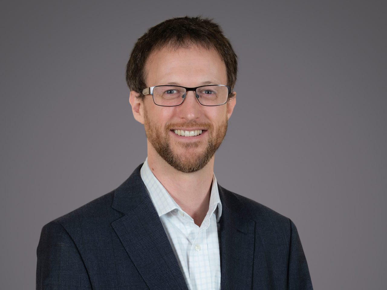 Person with short brown hair, trimmed beard and glasses, wearing a dark blazer over a light checked shirt, smiling against a plain grey background.