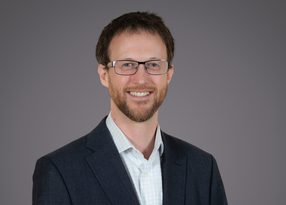 Person with short brown hair, trimmed beard and glasses, wearing a dark blazer over a light checked shirt, smiling against a plain grey background.