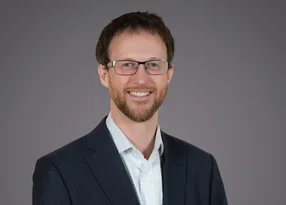 Person with short brown hair, trimmed beard and glasses, wearing a dark blazer over a light checked shirt, smiling against a plain grey background.