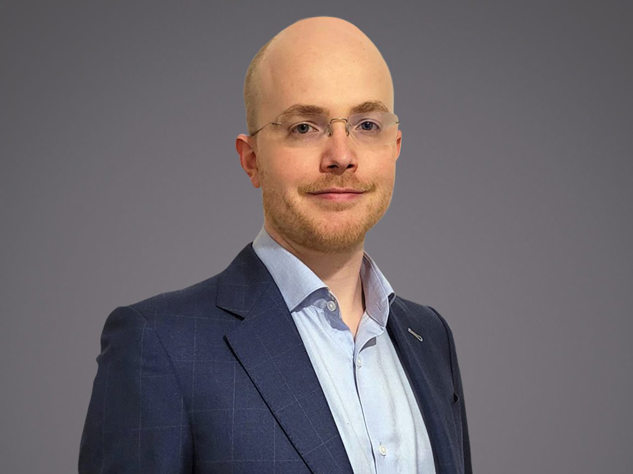 Head-and-shoulders portrait of a bald person wearing glasses, a light blue shirt and navy blazer, with a slight smile against a grey background.