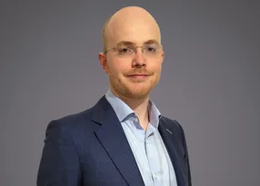Head-and-shoulders portrait of a bald person wearing glasses, a light blue shirt and navy blazer, with a slight smile against a grey background.