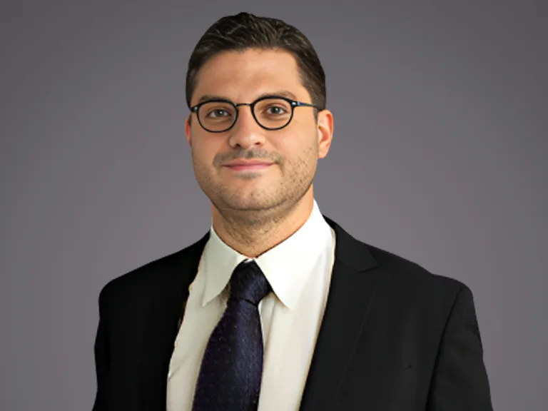 Individual with short dark hair and round glasses, wearing a black suit, white shirt and dark tie, against a grey gradient background.
